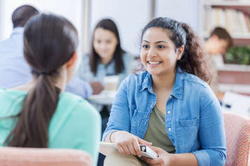 Teens sitting across from each other in chairs. One teen smiles at the other and holds a binder on her lap
